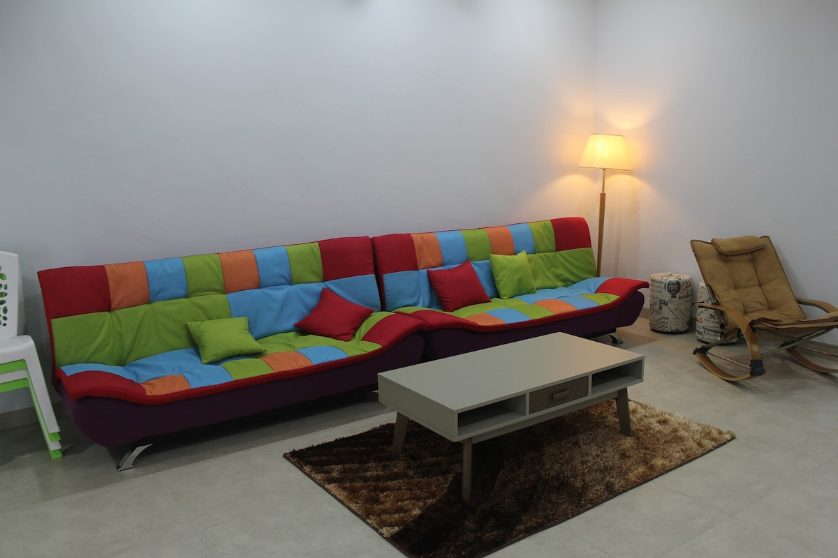 A colorful living area features a bold sofa with vibrant cushions in shades of red, green, and blue. A sleek, light-colored coffee table rests on a soft rug, while a warm lamp provides ambient lighting. A rocking chair is positioned to enhance relaxation.
