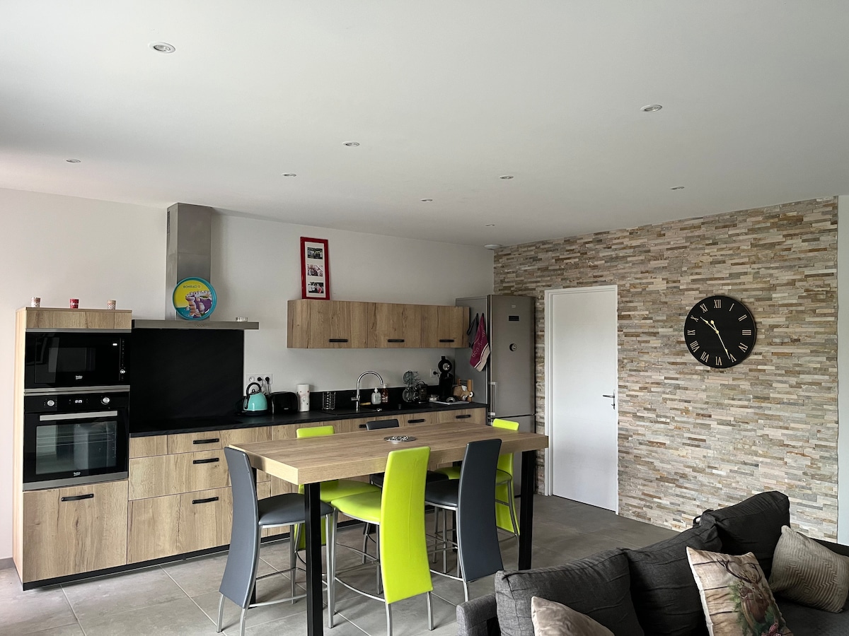 A modern kitchen features wooden cabinetry and stainless steel appliances. A large table with bright green chairs is positioned in the center, accompanied by a cozy gray sofa nearby. Natural light brightens the space, complemented by decorative stone wall accents and a wall clock.