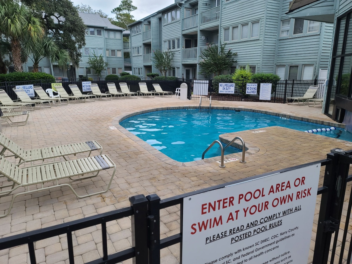 The pool area features a well-maintained circular pool surrounded by a spacious deck. Multiple lounge chairs are positioned around the pool, and clear signage is visible regarding safety rules. Lush greenery is present in the background, adding to the relaxed atmosphere.