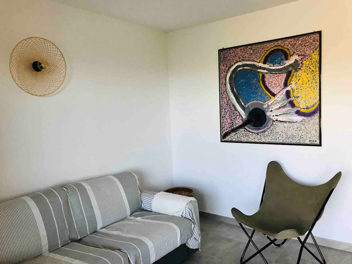 A modern living area is presented, featuring a striped sofa with a light throw draped over it. A bold abstract artwork adorns the wall, complemented by a unique, green folding chair in the corner. Natural light brightens the neutral-toned space.