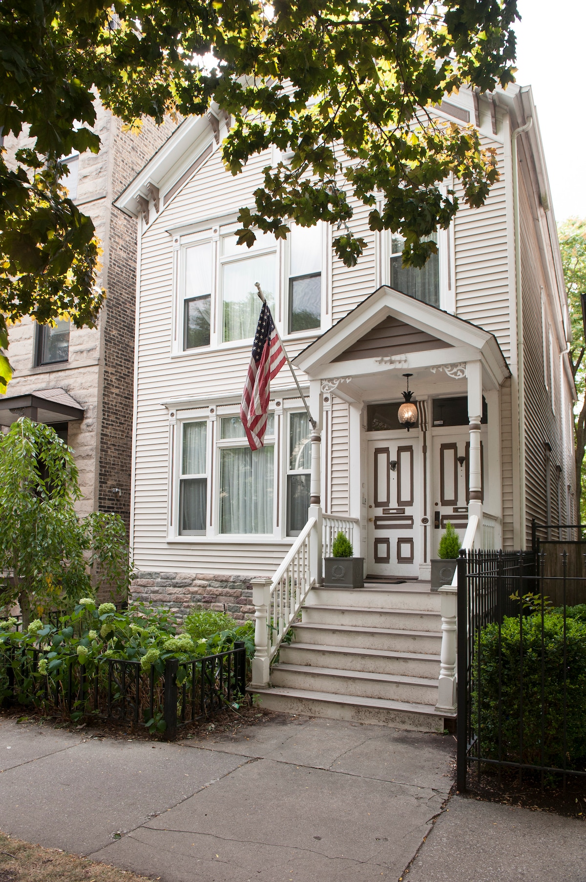 The exterior of a charming Victorian house is showcased, featuring a white facade, large windows, and a decorative entryway. An American flag hangs from the porch, surrounded by greenery and a well-maintained pathway leading to the entrance.