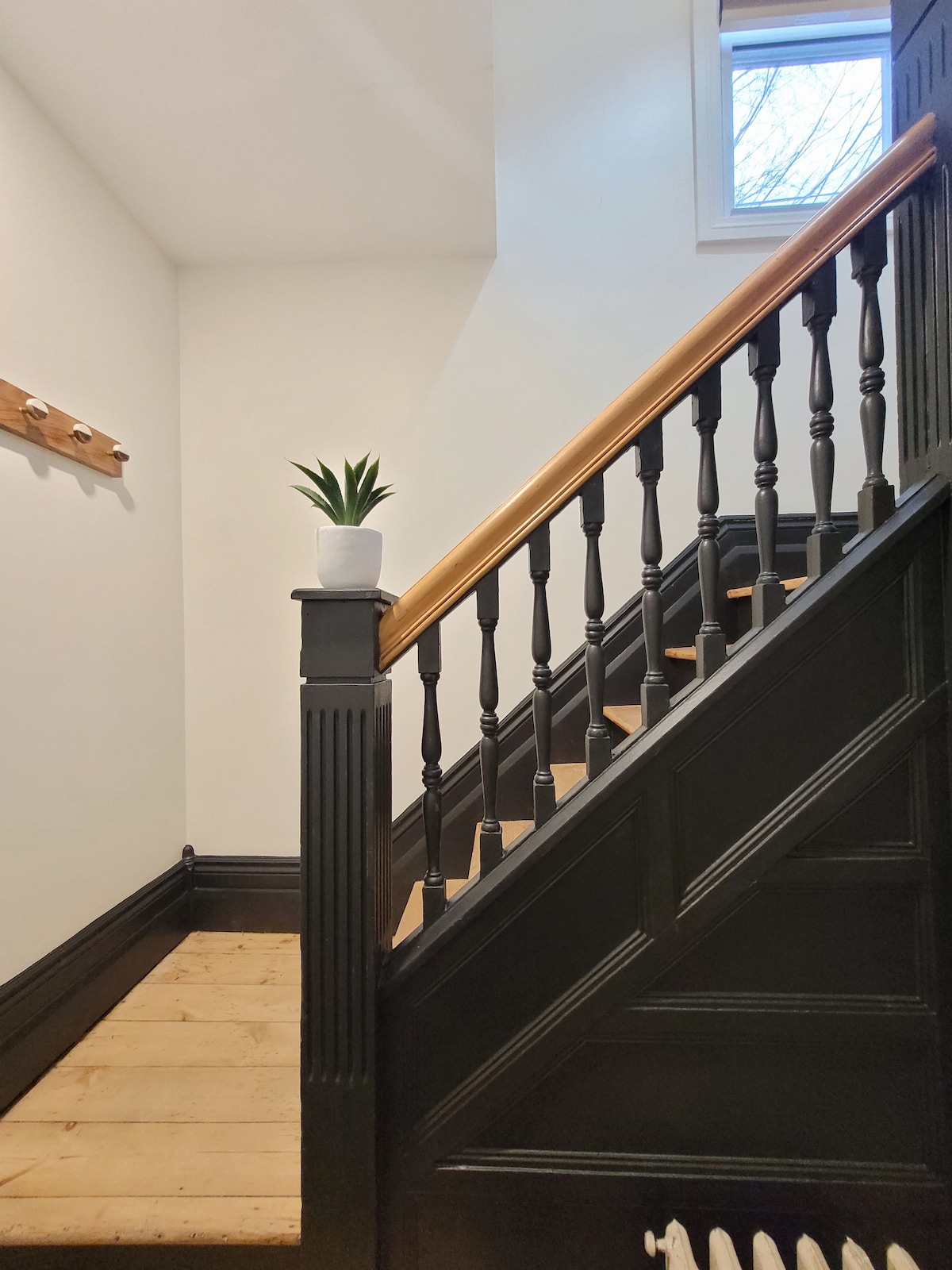 A well-lit staircase features wooden steps leading up, flanked by a black railing with decorative spindles. A potted plant sits on a ledge at the top, and natural light filters in through a window, enhancing the clean and inviting space.