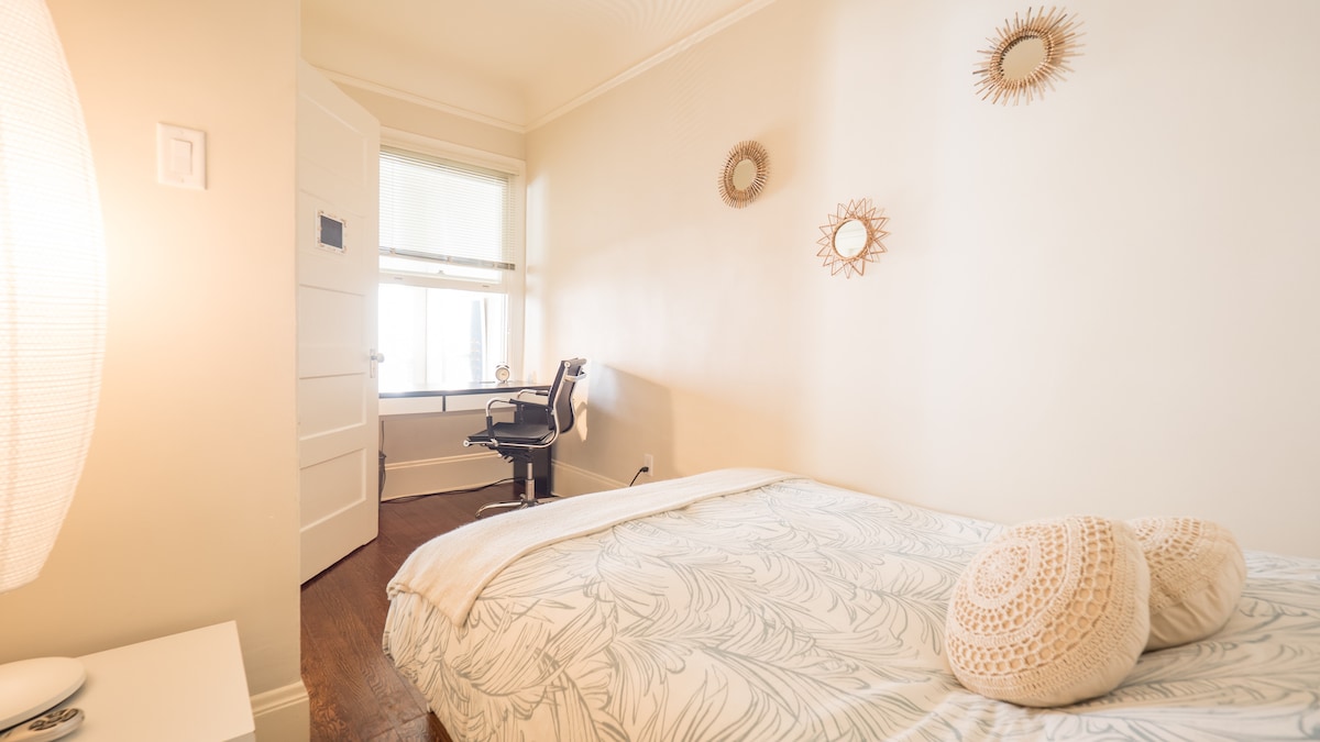 A small room features a comfortable full-size bed with soft bed linens. A desk is positioned near a window, enhancing the light in the space. Three decorative mirrors adorn the wall, complementing a warm and minimalistic decor.