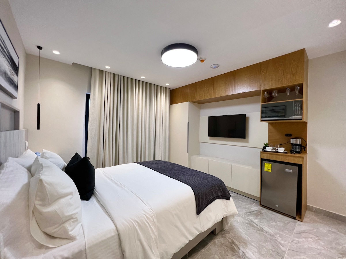 A modern bedroom features a spacious bed with white linens and a dark throw blanket. A television is mounted on a light wood accent wall, above a sleek cabinet. A kitchenette with a mini fridge and coffee maker is present, and soft lighting is provided by a ceiling fixture.