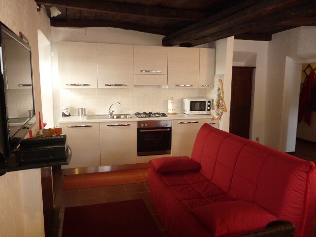 The compact kitchen area includes modern cabinetry and appliances, such as a gas stove, microwave, and refrigerator. A red sofa bed is positioned in the foreground, offering a pop of color. Warm lighting enhances the cozy atmosphere created by the wooden beams overhead.