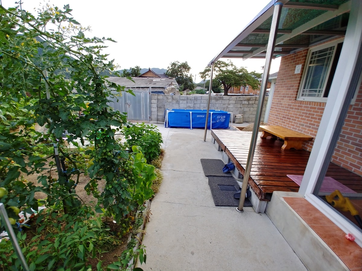 A pathway lined with greenery leads to a wooden seating area under a covered porch. A blue above-ground pool is visible in the background, bordered by a stone wall. Adjacent to the pathway, a garden area can be seen, enhancing the outdoor setting.