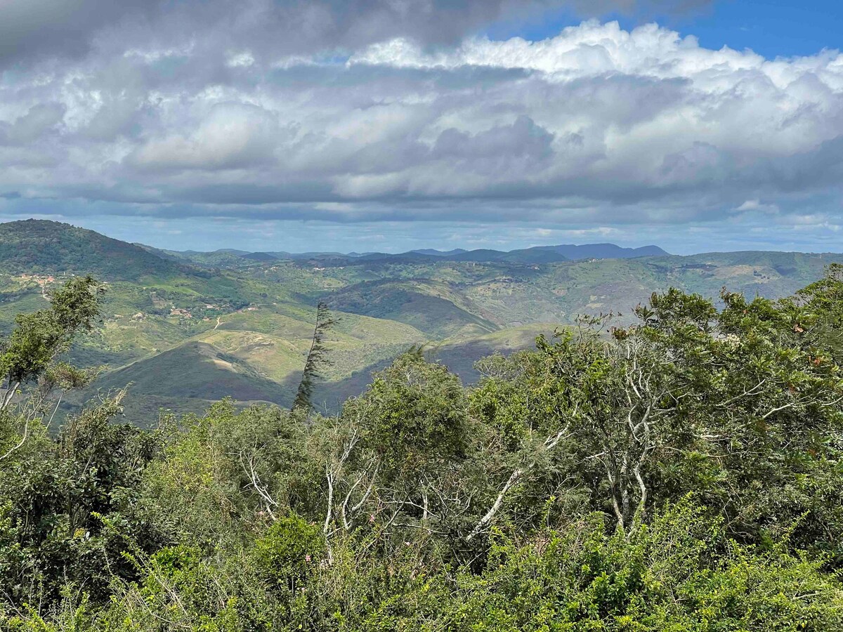 A panoramic view of rolling green hills and valleys is presented, with a mix of trees and shrubs in the foreground. The landscape is partially covered by scattered clouds, allowing sunlight to illuminate different areas of this tranquil natural setting.