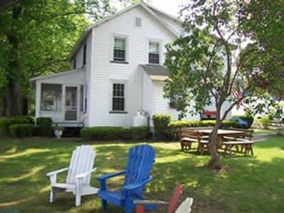 The exterior of a two-story lake house is depicted, featuring a white façade and surrounded by green landscaping. Adirondack chairs in blue and white are placed on the lawn. A picnic table is visible nearby, under the shade of trees, contributing to the outdoor gathering space.