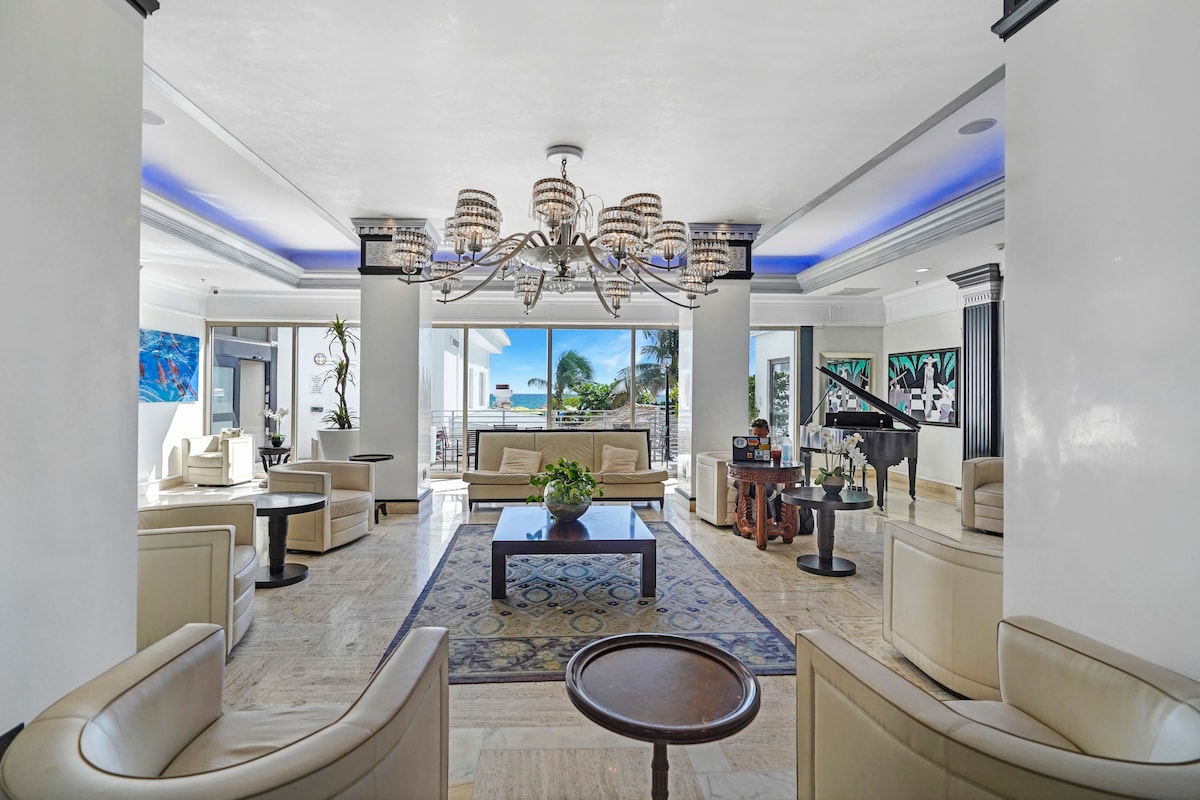 A spacious lobby area features elegant seating arrangements with light-colored sofas and armchairs surrounding a central area rug. Large windows provide views of the outdoor space, while a chandelier illuminates the room. Potted plants add a touch of greenery to the inviting atmosphere.