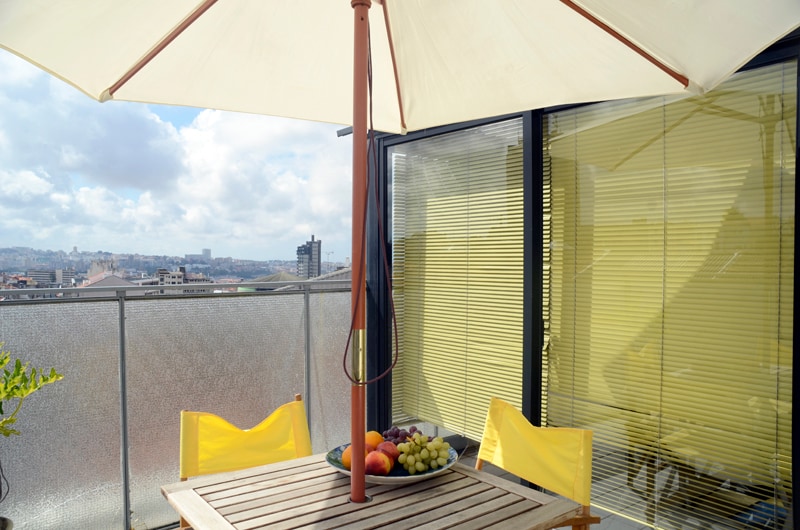 A terrace table made of wood is accompanied by two yellow chairs and shaded by a large umbrella. A bowl filled with various fruits is placed center table. The background features views of the city skyline, with bright skies and distant buildings visible.