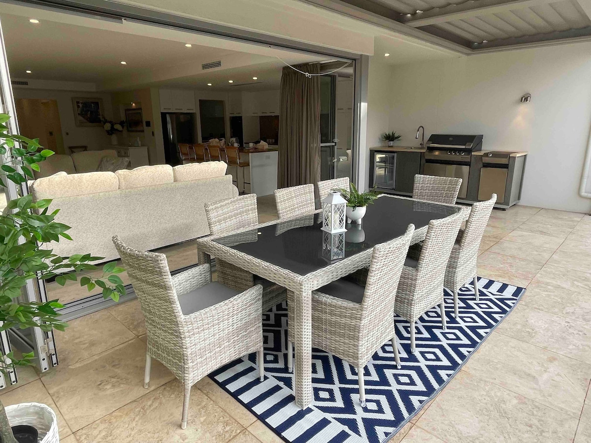 An outdoor dining area is presented with a large rectangular table surrounded by six wicker chairs. A decorative rug enhances the space, and a gas BBQ is visible in the background. Sliding glass doors provide access to the adjoining indoor area.
