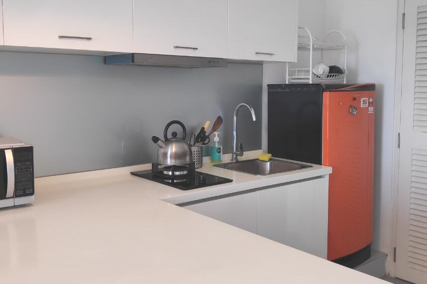 A modern kitchen features sleek cabinetry and a clean countertop. Essential appliances include a microwave, a kettle, and a vibrant orange refrigerator. A stainless steel sink is positioned near the stovetop, showcasing a minimalist and functional design.