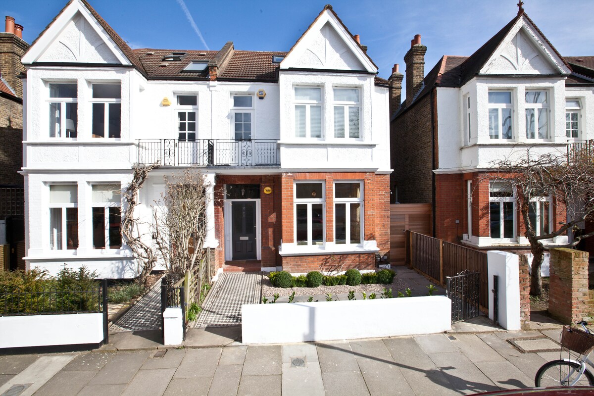 A charming family home is showcased, distinguished by its white and red brick facade. Large rectangular windows allow natural light to fill the interior, and a welcoming front pathway leads to the entrance. A neatly trimmed hedge and decorative plants frame the property.