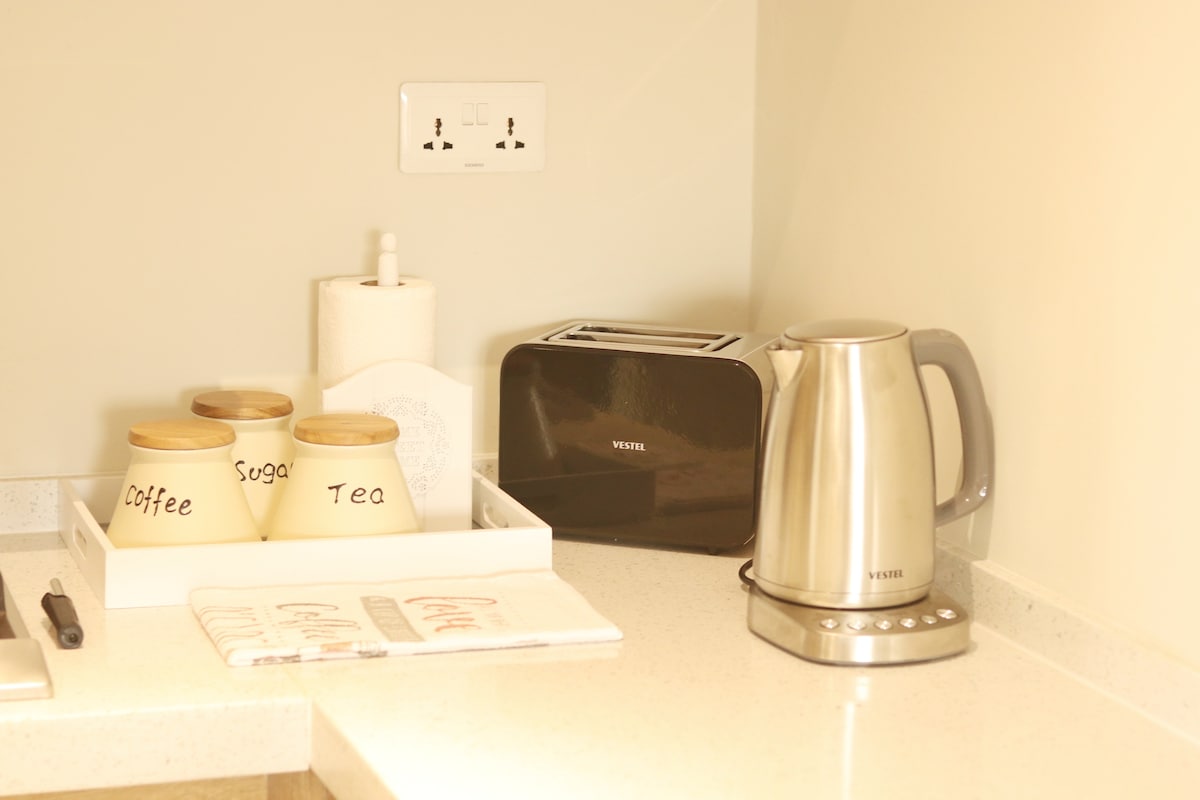 A modern kitchen countertop is displayed, featuring a stainless steel kettle and a sleek toaster. A tray holds labeled jars containing coffee, sugar, and tea. A roll of paper towels is positioned beside the jars, contributing to the functionality of the space.