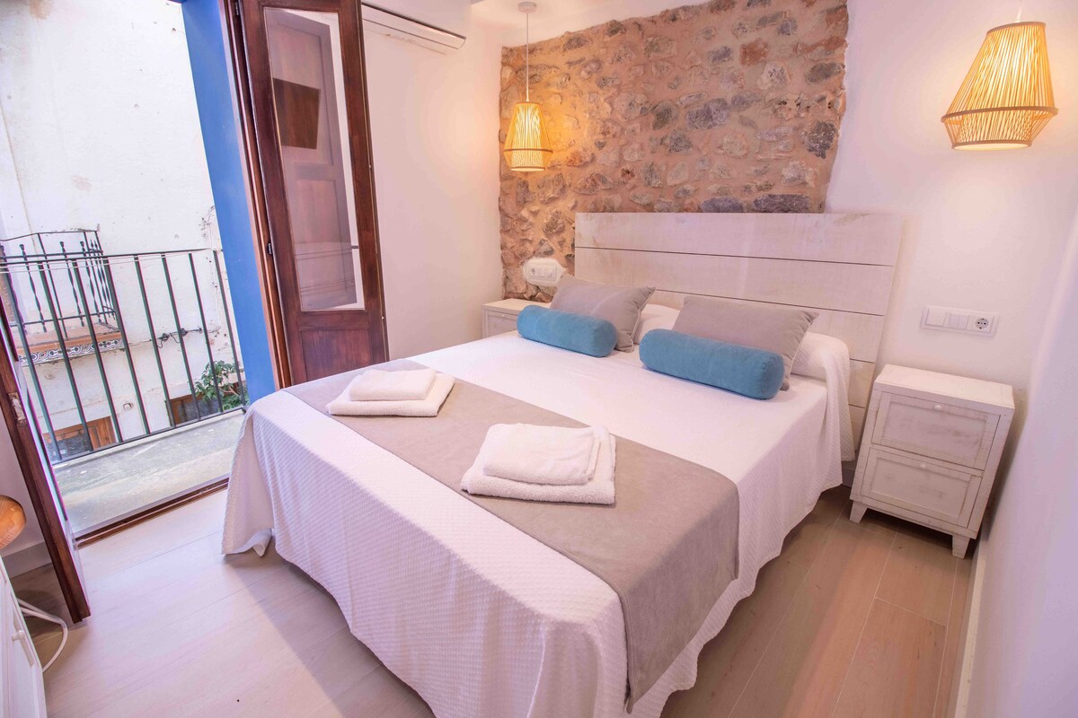 A bright sleeping area features a spacious bed adorned with blue-accented pillows and neatly arranged towels. Textured stone walls add character, while wooden light fixtures create a warm glow. A balcony with a railing is visible through the open door, inviting natural light into the room.