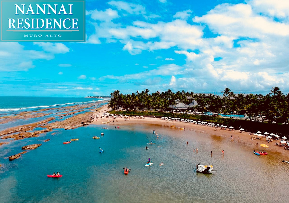 An aerial view captures the tranquil shoreline of Muro Alto, showcasing a crescent-shaped beach with gentle waves. Sunbathers and kayakers enjoy the water as palm trees line the coast, providing shade. Umbrellas and lounge chairs are visible along the sandy beach.
