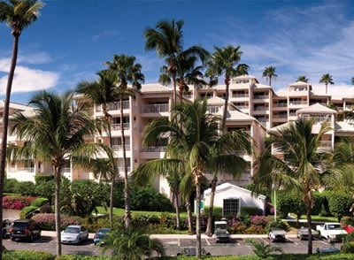 A multi-story resort building is surrounded by lush palm trees and vibrant tropical landscaping. The architecture features a light-colored facade, and parked cars are visible in the foreground, highlighting the welcoming atmosphere of the resort.