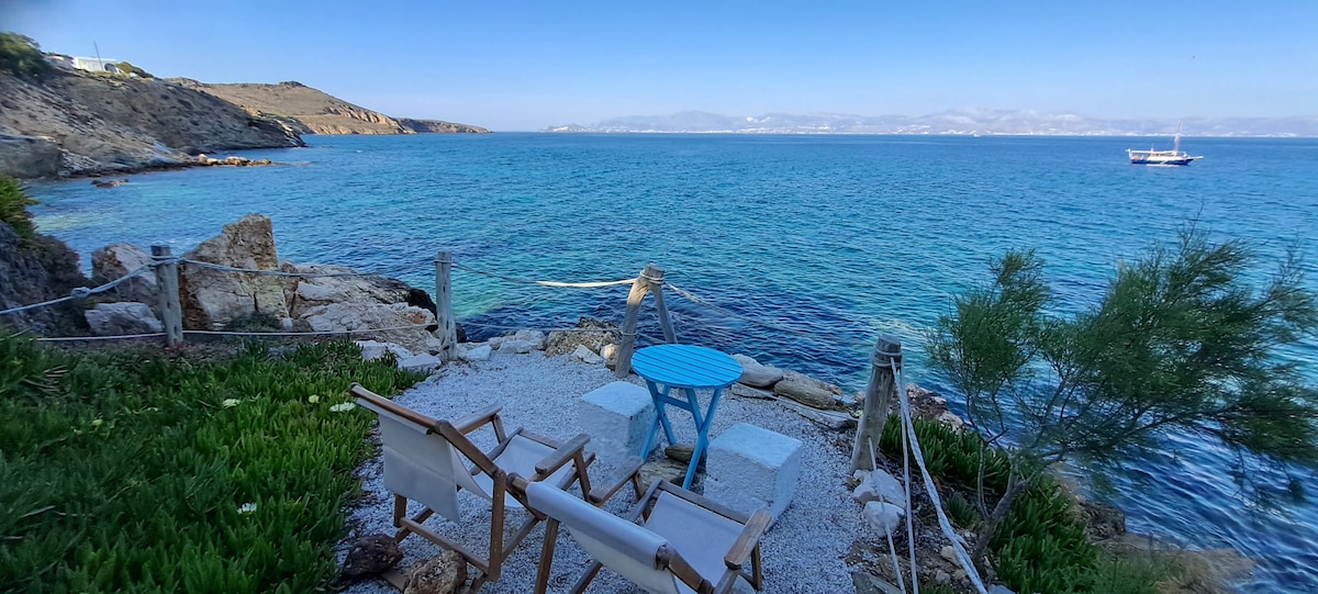 A serene waterfront space is presented, featuring a small round blue table with two white chairs. The crystal-clear turquoise water flows gently towards rocky shores, while distant hills and a boat can be seen across the horizon under a clear blue sky.