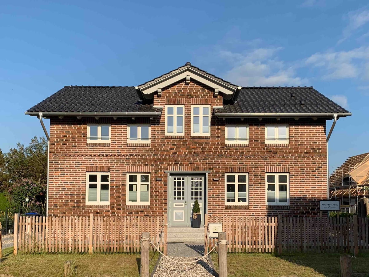 A two-story brick building features a sloped roof and multiple windows on the façade, allowing for natural light. A fenced pathway leads to the front door, bordered by well-maintained grass. The overall architectural style reflects a blend of modern and traditional elements.
