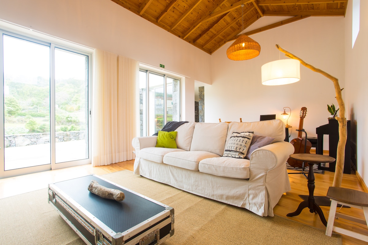 A spacious living area features a comfortable white sofa adorned with a colorful pillow, set against a backdrop of large glass doors that open to the outdoors. Natural light floods the space, highlighting wooden beams on the ceiling and a stylish pendant light overhead.
