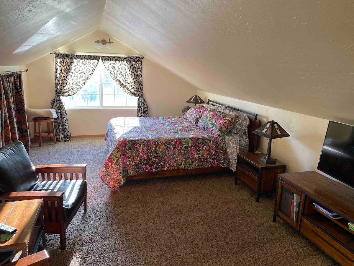 A spacious loft area features a king-size bed with vibrant floral bedding. A comfortable seating arrangement includes a dark leather chair and wooden side table. Natural light fills the space through a large window adorned with patterned curtains, and a television is positioned on the opposite wall.