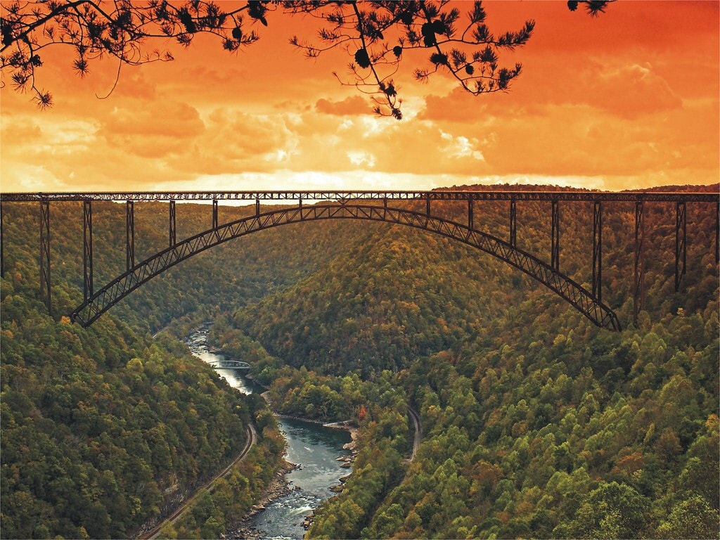 A striking view of the New River Gorge Bridge spans across a vibrant valley, surrounded by dense greenery and autumn foliage. The bridge's arching structure is set against an orange and golden sky, reflecting the natural beauty of the landscape below, where the river winds through the gorge.