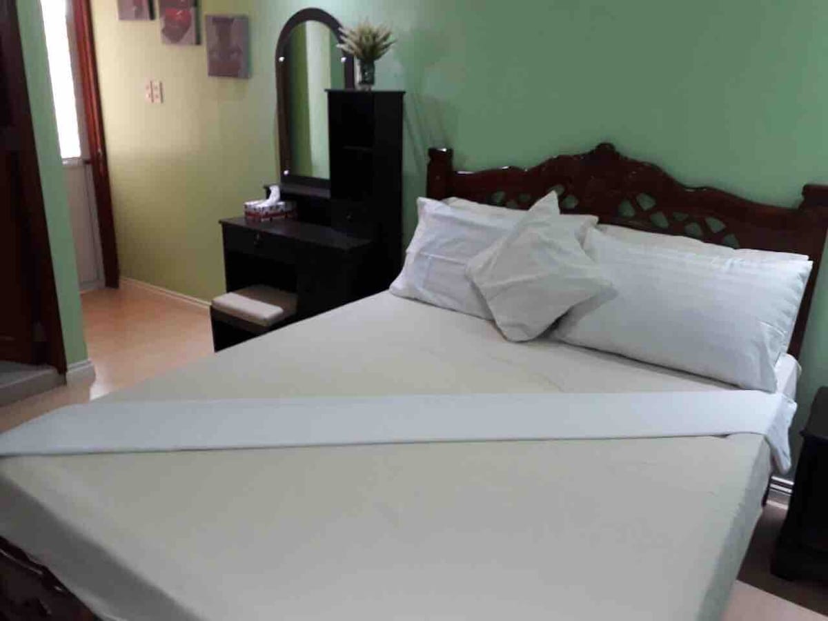 A spacious sleeping area features a Full Double bed with neatly arranged linens and decorative pillows. A black wooden dresser with a mirror and a small seating area are positioned nearby, complemented by softly painted green walls that create a calming atmosphere.