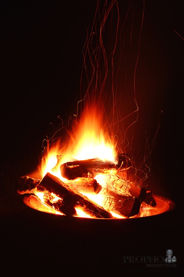 A glowing fire is contained within a circular pit, with logs arranged strategically to enhance the flames. The orange and yellow hues contrast against a dark background, creating a cozy ambiance. Sparks are seen dancing upwards, adding a dynamic element to the scene.