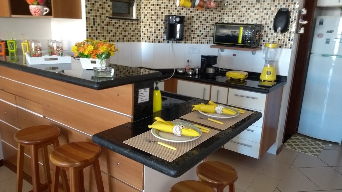 A modern kitchen with wooden cabinetry and a mosaic tiled backsplash is featured. A dining table set for four is displayed, with yellow napkins and stylish table settings. A microwave, coffee maker, and blender are visible, alongside a refrigerator and stove.