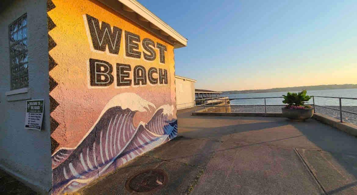 A vibrant mural depicting waves is displayed on the side of a building, with 'West Beach' prominently painted in bold letters. The sun sets in the background, casting a warm glow over the pathway that leads toward the shoreline.