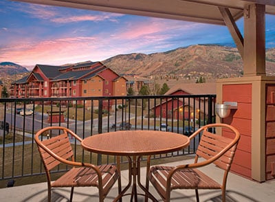 A balcony features a round table and two chairs, offering a view of the scenic mountains at sunset. The charming landscape includes resort buildings and lush greenery, set against a colorful sky.