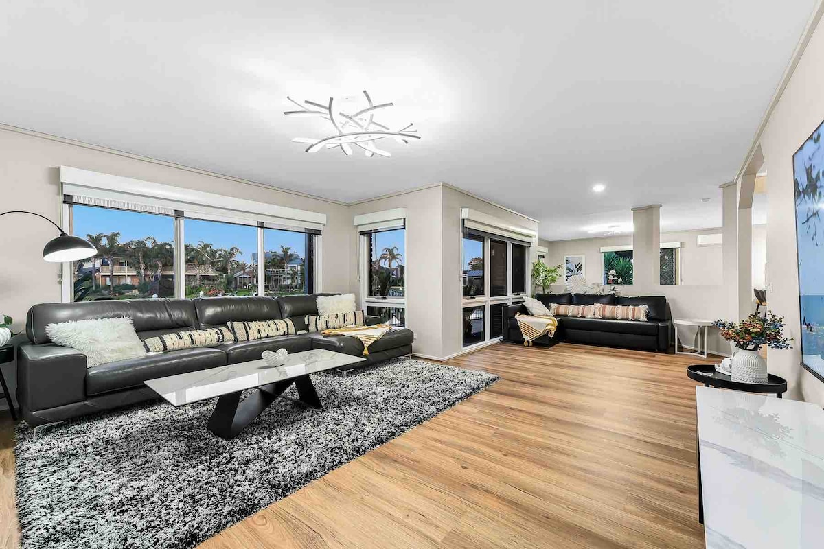 A modern living area features dark leather sofas, accompanied by a glass coffee table on a large, textured rug. Sleek lighting adorns the ceiling, while large windows allow natural light to fill the space. Decorative plants and a minimalist art piece enhance the room's ambiance.