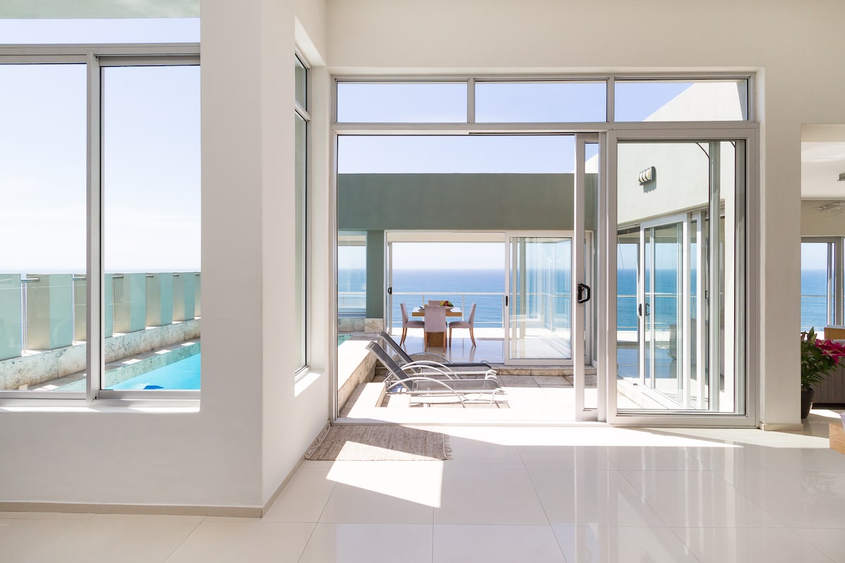 A spacious living area is framed by large glass doors, offering stunning ocean views. Sun loungers are positioned near the pool, providing a relaxing spot. A light-colored dining table is visible in the background, enhancing the open, airy feel of the space.