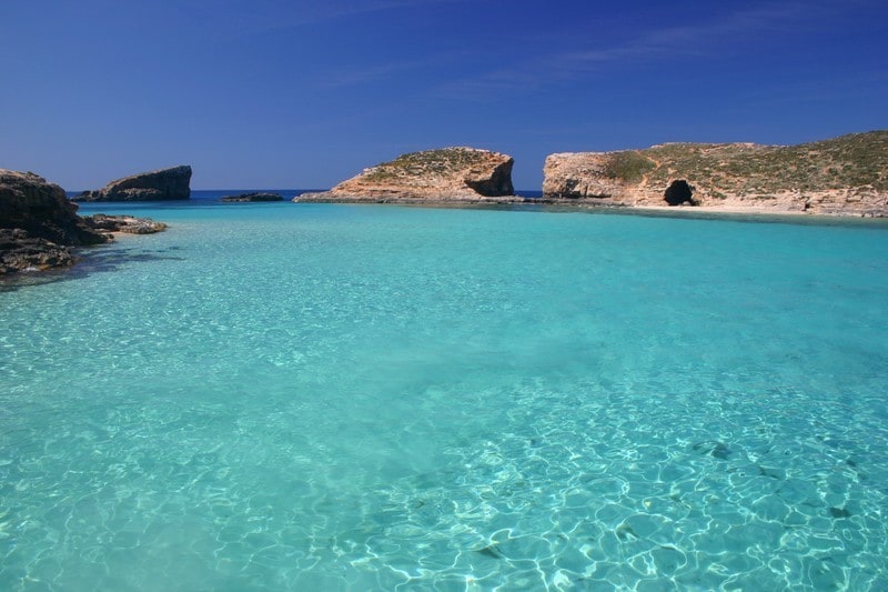 A serene view of crystal-clear turquoise water is depicted, showcasing the gentle ripples and sandy seabed beneath. In the distance, rocky islands rise from the tranquil sea, highlighted against a bright blue sky with a few wispy clouds.