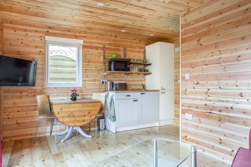 A warm wood interior defines the kitchen and dining area, featuring a round wooden table paired with four chairs. A kitchenette with modern appliances is present, along with open shelving displaying dishware. A television is mounted on the wall, and a window allows natural light to enter.