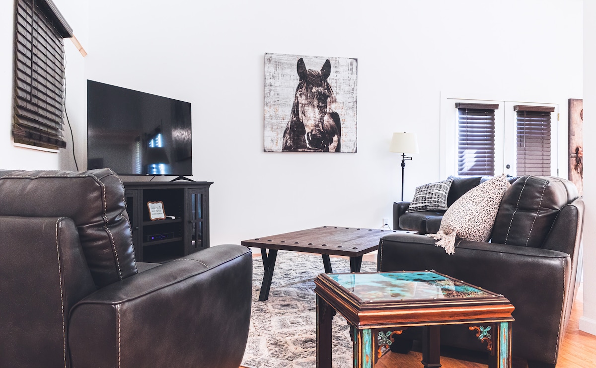 A welcoming living area features two dark leather sofas facing a low wooden coffee table, complemented by a decorative table. A large television sits on a dark console against a white wall, while a horse-themed artwork adds a rustic touch. Natural light enters through partially covered windows.
