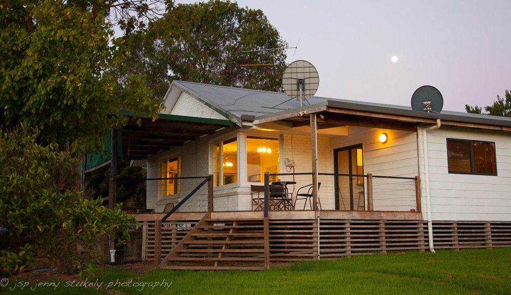 A family farmhouse features a covered porch with a small outdoor dining set overlooking lush greenery. Satellite dishes are mounted on the roof, with the moon visible in the evening sky. The light from the interior complements the tranquil surroundings.