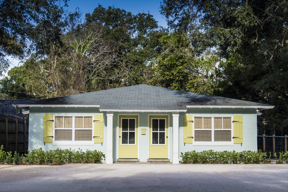 A charming duplex is shown with a light blue exterior and yellow shutters framing two inviting front doors. The property is surrounded by lush greenery and a gravel pathway, creating a welcoming environment. Tall trees provide shade and enhance the setting.