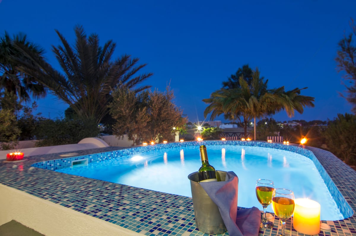 A plunge pool featuring hydromassage facilities is illuminated by soft lighting in the evening. Nearby, a bottle of wine rests in an ice bucket, accompanied by two glasses filled with a golden beverage. Tropical plants surround the area, adding to the serene environment.