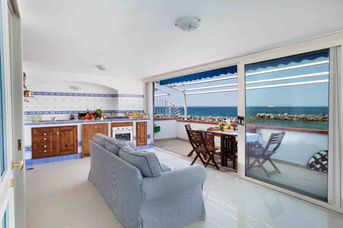 An airy living space includes a comfortable sofa and a dining area with wooden chairs positioned near large glass doors. A well-equipped kitchen features wooden cabinetry and modern appliances, providing an inviting view of the sea through the spacious windows.