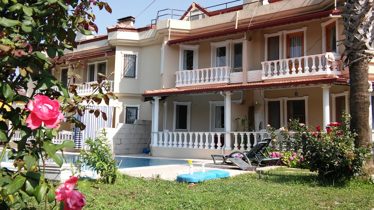 A three-story villa is showcased, featuring a well-maintained garden with blooming roses. The facade includes balconies with decorative railings. A private pool area is visible, complemented by sun loungers and a backdrop of vibrant greenery.