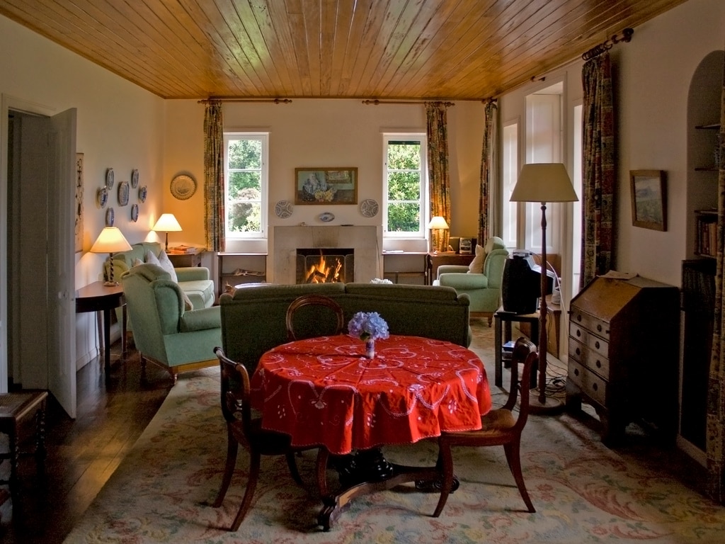 A spacious living area is presented, featuring two comfortable armchairs and a green sofa arranged around a central round table draped with a red tablecloth. Natural light enters through two large windows, illuminating the space, which includes a fireplace and warm wooden accents.