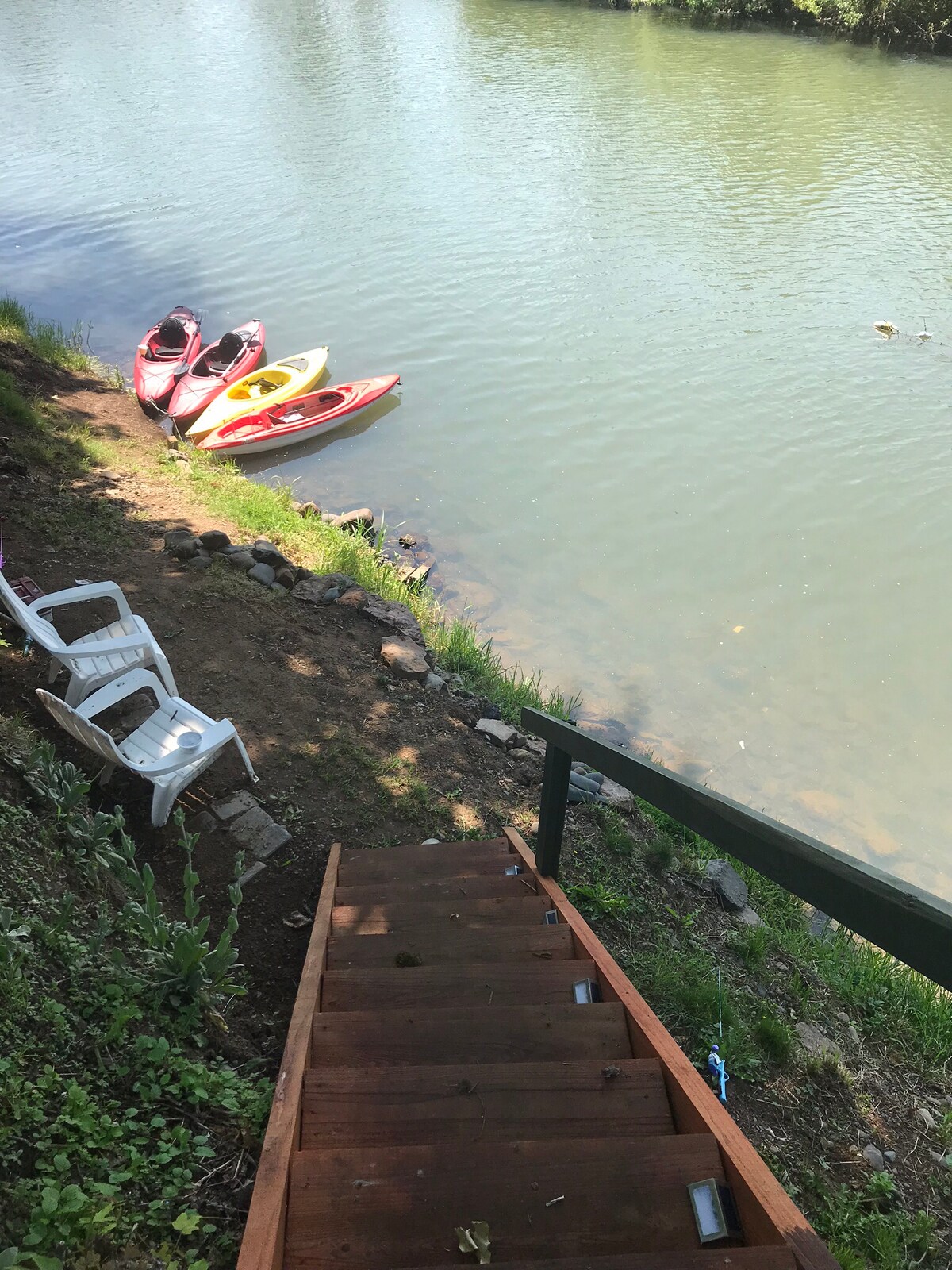 A view of the riverbank features two kayaks resting by the water's edge, one red and one yellow. A set of wooden steps leads down to the water, with two white chairs positioned nearby for relaxation. Lush greenery surrounds the area, enhancing the natural setting.