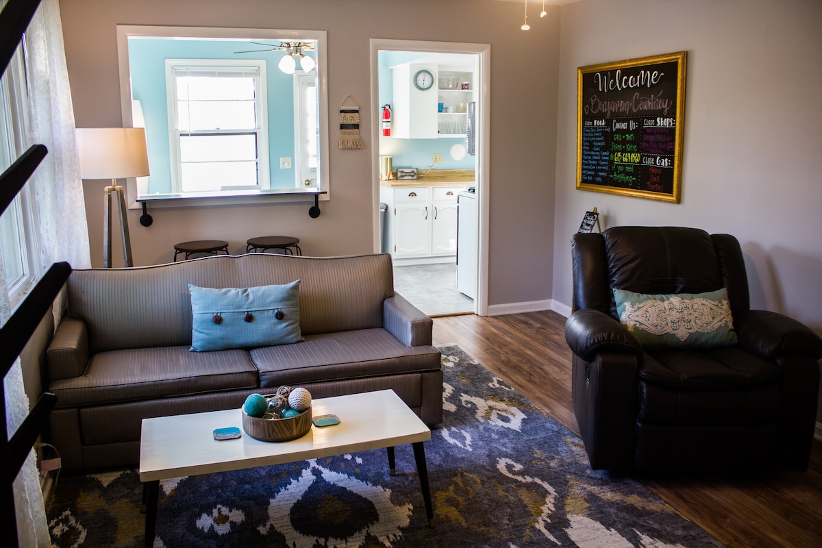 The living area features a comfortable sofa adorned with decorative pillows, a coffee table with decorative items, and a recliner. Natural light enters through large windows, illuminating the space. An open entry leads to a bright kitchen, creating an inviting atmosphere.