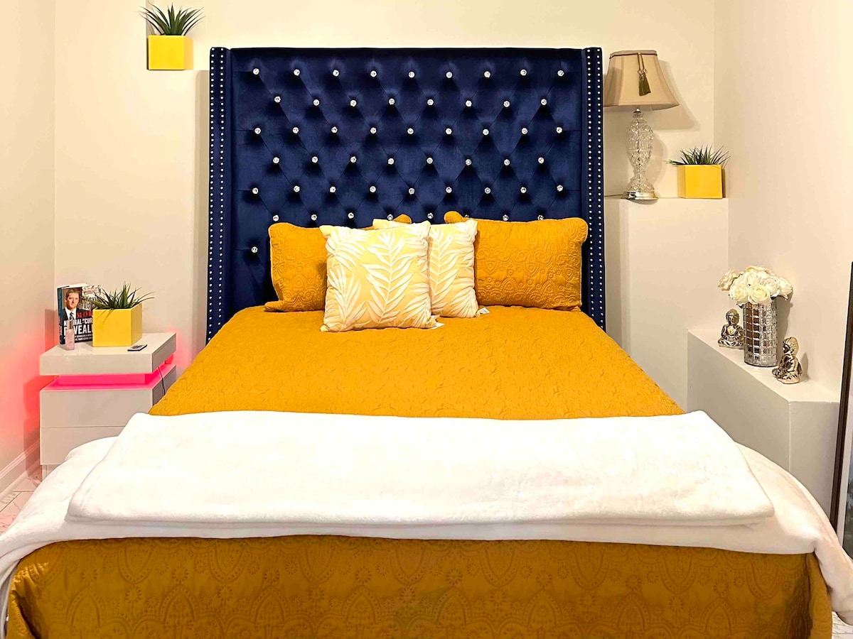 A stylish bedroom features a deep blue tufted headboard adorned with decorative studs. An inviting golden bedspread covers the mattress, layered with white and patterned throw pillows. Nightstands with yellow planters and lamps sit on either side, contributing to the room's modern design.