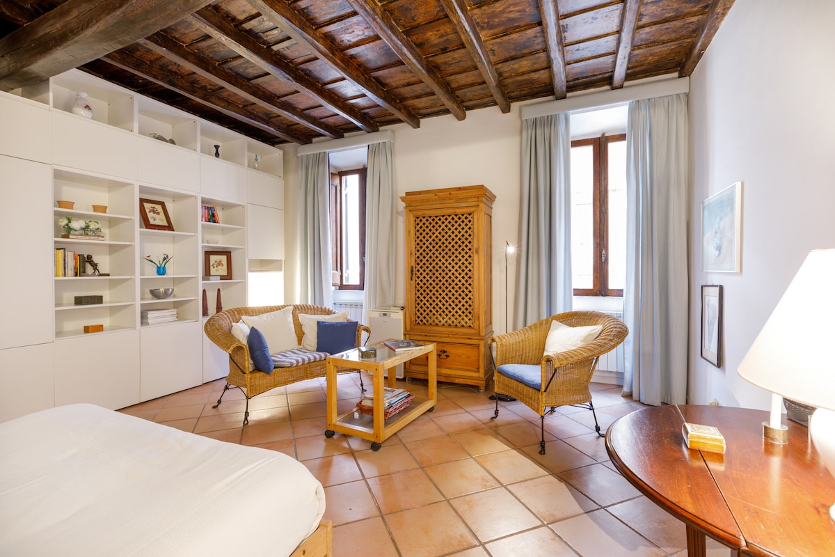 A bright bedroom features exposed-beam ceilings and two windows framed by soft curtains. A wicker seating area with a wooden table is positioned across from a spacious bed, while built-in shelving provides storage and decoration. A warm light from a lamp adds a cozy touch.