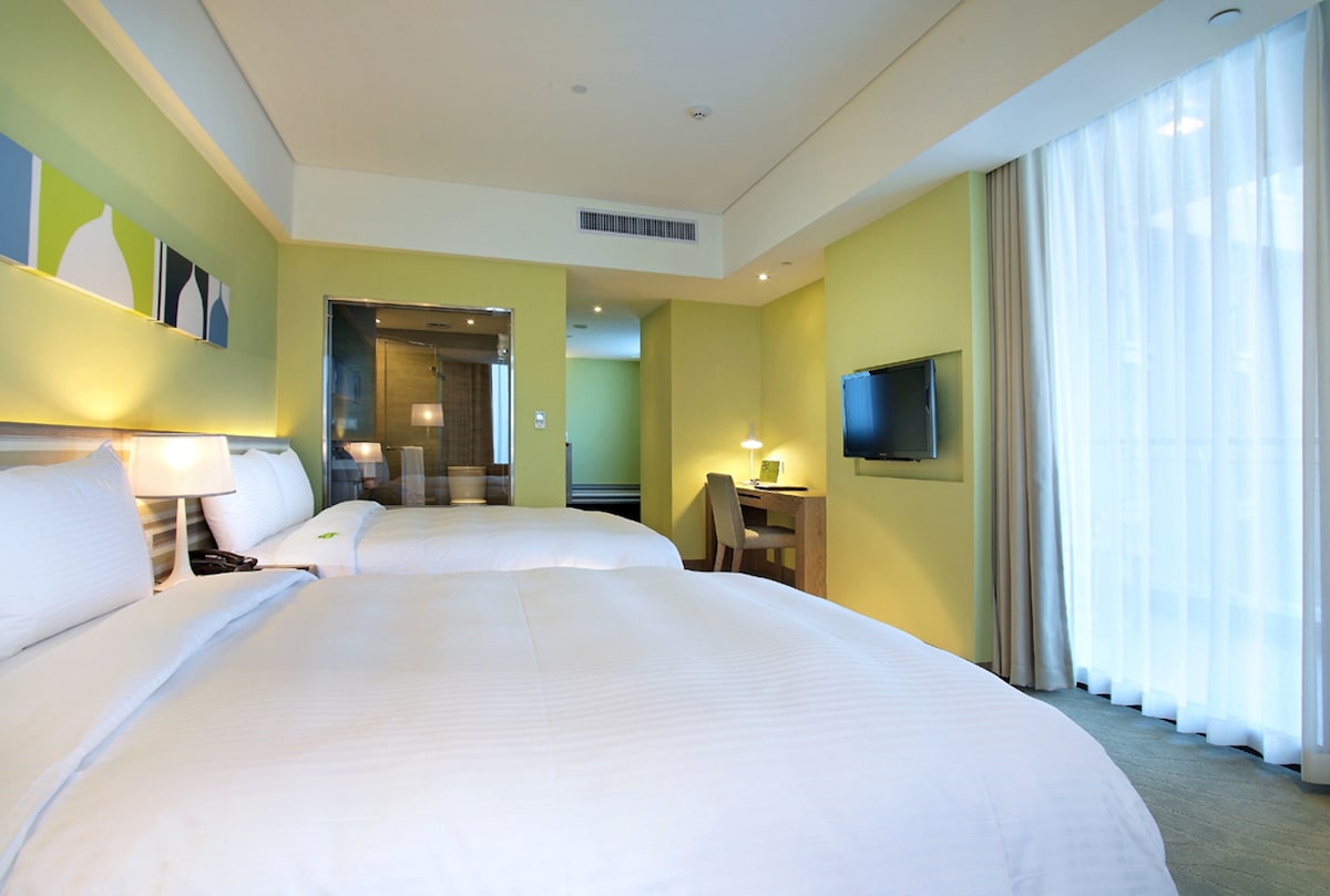 A well-lit guest room features two spacious double beds with crisp white linens. Soft green walls and modern artwork complement the ambiance. A flat-screen TV is mounted on the wall, while a desk and chair are positioned nearby. Large windows allow natural light to flood the space.