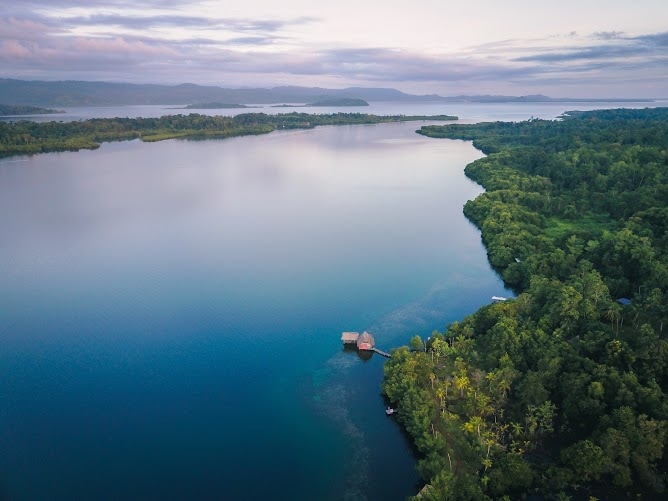 An aerial view captures a serene waterfront landscape, where lush greenery lines the shoreline and calm waters extend towards distant hills. A small dock with a rustic structure is visible, inviting guests to explore the peaceful surroundings at sunset.