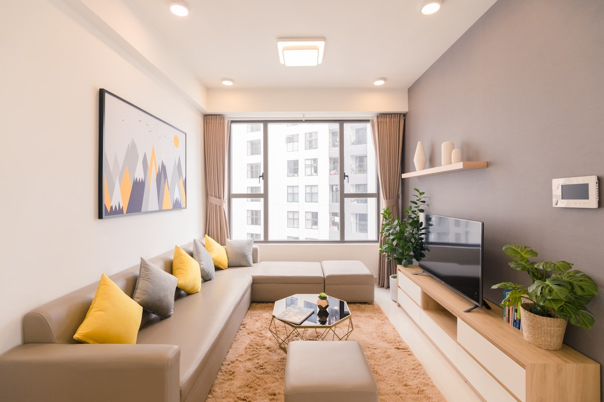 The living room features a sectional sofa adorned with colorful cushions, arranged invitingly around a coffee table. A large window allows natural light to fill the space, complemented by neutral walls and a modern entertainment unit housing a TV. Decorative elements, including plants, add a fresh touch.