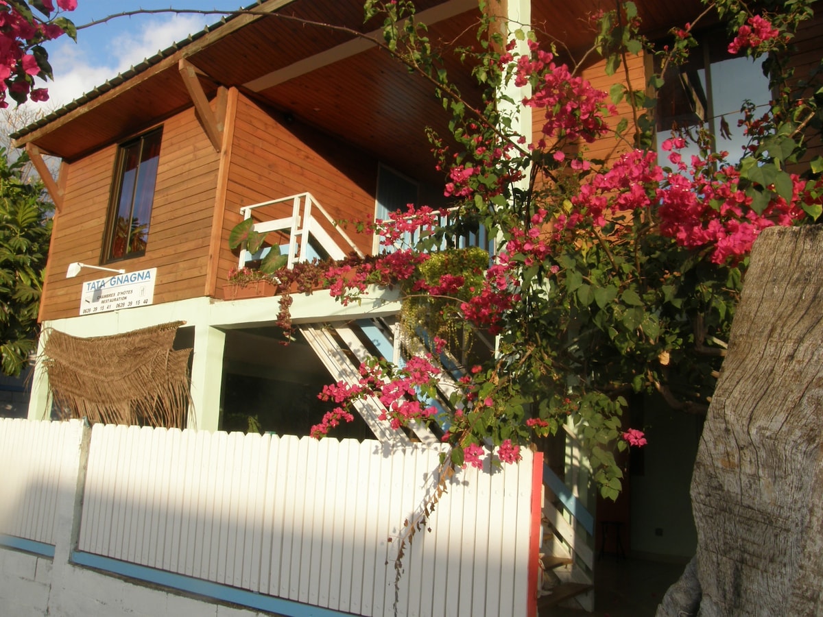 A wooden exterior features vibrant bougainvillea cascading over a balcony. Stairs lead to the upper level, where large windows invite natural light. A white picket fence surrounds the property, creating a charming boundary while showcasing the unique architecture.